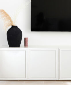 Black vase with tall dried pampas grass on a white cabinet beside a wall-mounted TV; a red speckled candle sits nearby.