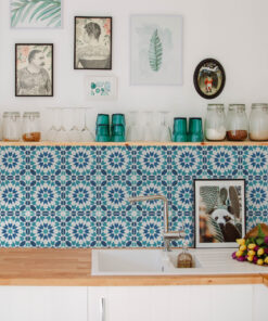 White kitchen counter scene with glass jars, teal cups, and a faucet set against a blue geometric tile wall with framed art above.