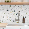 02 copy | Casadart | Vinis Autocolantes, Papel de Parede e Painéis Decorativos Kitchen sink area with terrazzo-pattern backsplash, chrome faucet, brown soap dispenser, and cutting board on white counter.