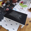01 | Casadart | Vinis Autocolantes, Papel de Parede e Painéis Decorativos Play area for children with stuffed animals, crayons, a drawing, and a green toy bin on a star-pattern rug.