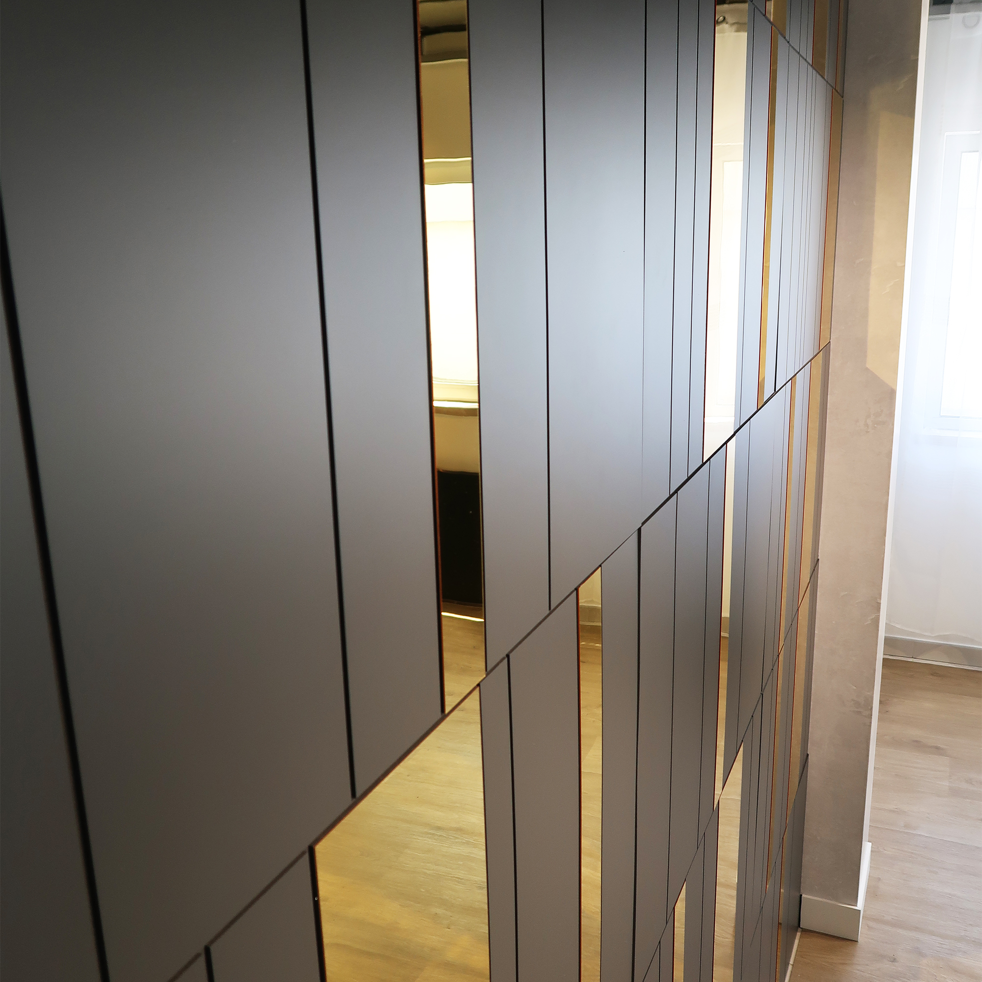 IMG_3729 copy | Casadart | Vinis Autocolantes, Papel de Parede e Painéis Decorativos Interior view of a modern wall with tall, dark grey vertical panels and narrow gold-tinted glass strips, a wooden floor visible at the bottom.