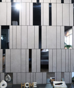 Modern interior showroom display: beige tiled wall with vertical black gaps, glass openings revealing decor like white vases, a plant, magazines, and the word 'HOME' on the shelf.