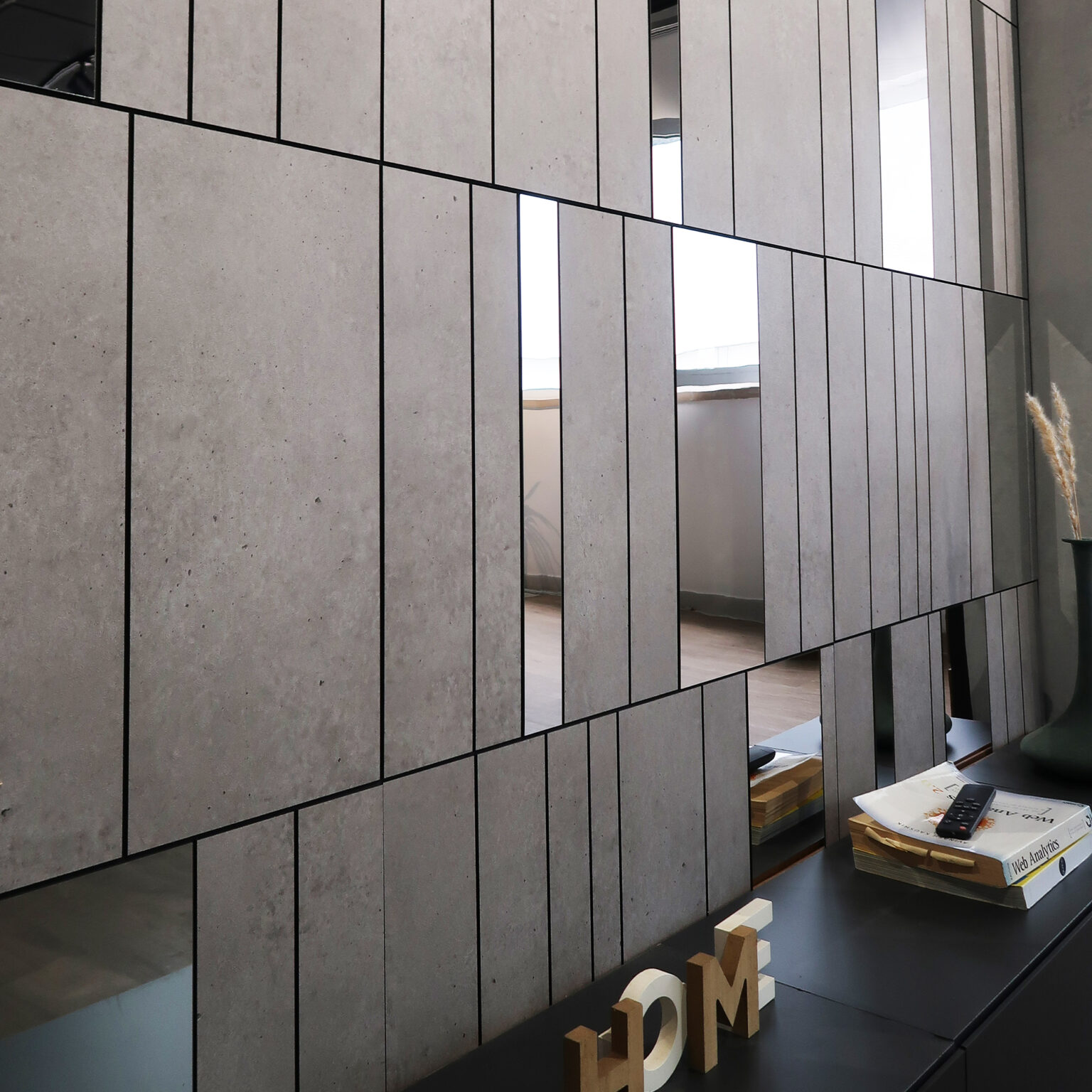 Modern interior wall of concrete panels with black grout lines and diagonal seam, plus a desk with magazines and wooden letter blocks seen on the right edge.
