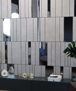 Showroom display with a gray paneled wall and vertical slits, a black cabinet, white ceramic vases, a wooden 'HOME' sign, blue cloth, and a sofa visible through the openings.