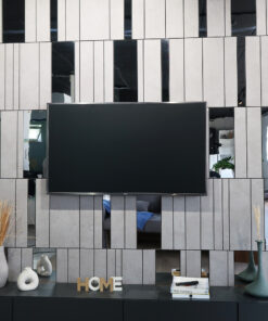 Mounted flat-screen TV on a gray tiled wall with white vases, books, green plants, and a decorative 'HOME' sign on the shelf.] ,