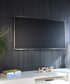 Large flat-screen TV mounted on a dark paneled wall with a green plant to the left and wooden 'HOME' letters on a shelf below.