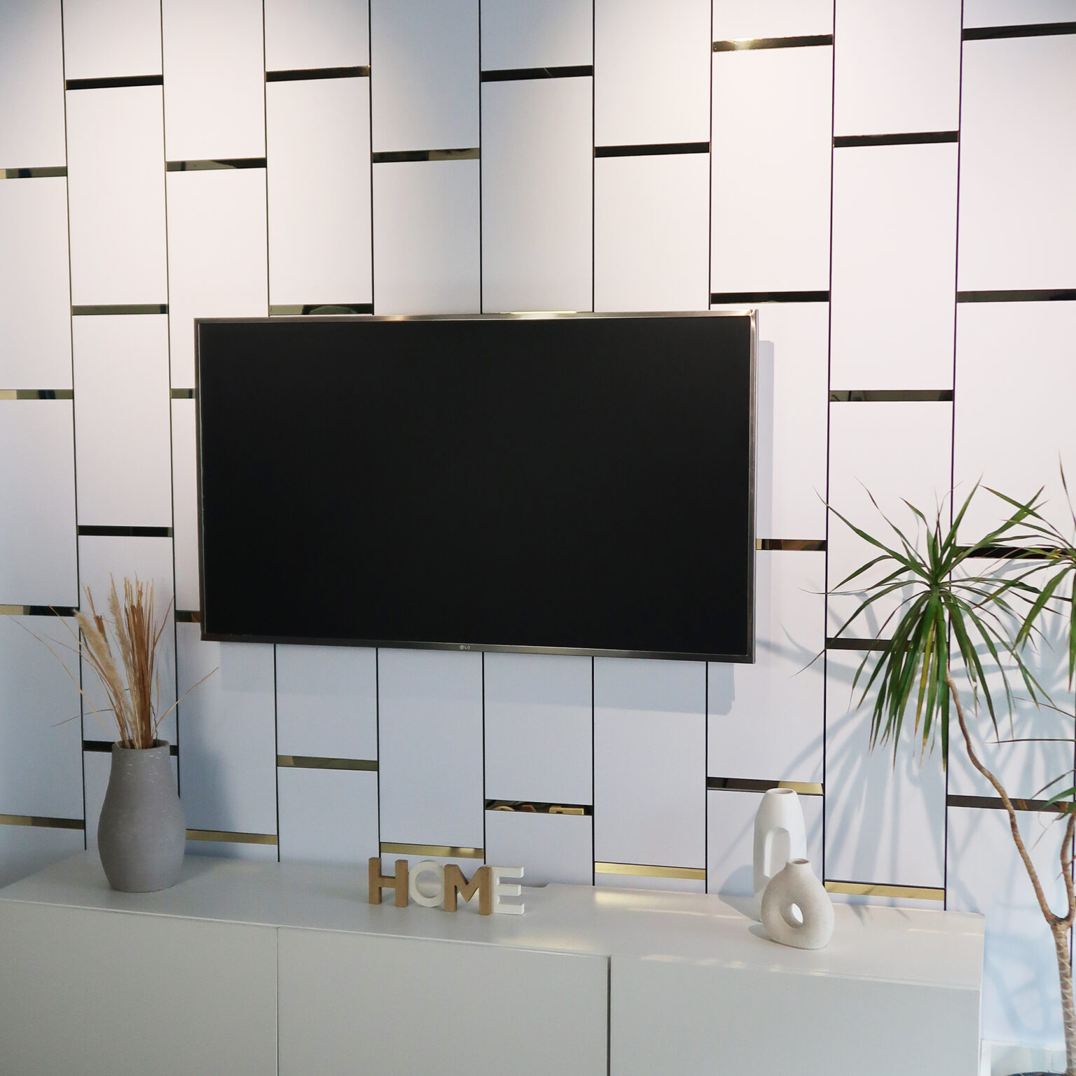 White paneled wall with gold accents, a mounted flat-screen TV, and decorative vases and a plant on a shelf below.