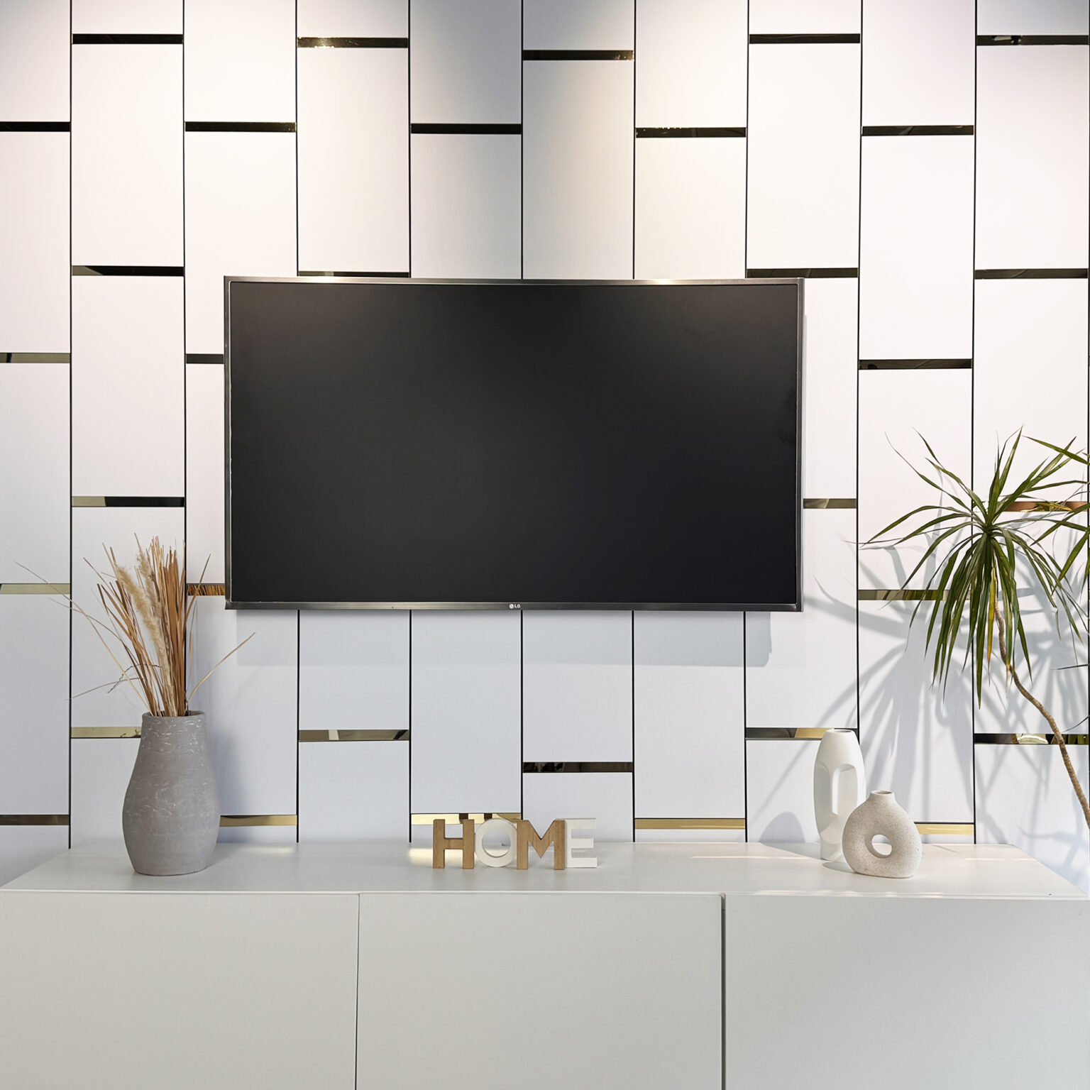 Modern living room with a wall of white panels accented by thin gold strips, a large flat-screen TV, and vases of dried grasses on a white cabinet.