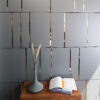 | Casadart | Vinis Autocolantes, Papel de Parede e Painéis Decorativos Wooden table with a tall gray vase holding dried grass and an open magazine, set against a gray slatted wall with gold gaps.