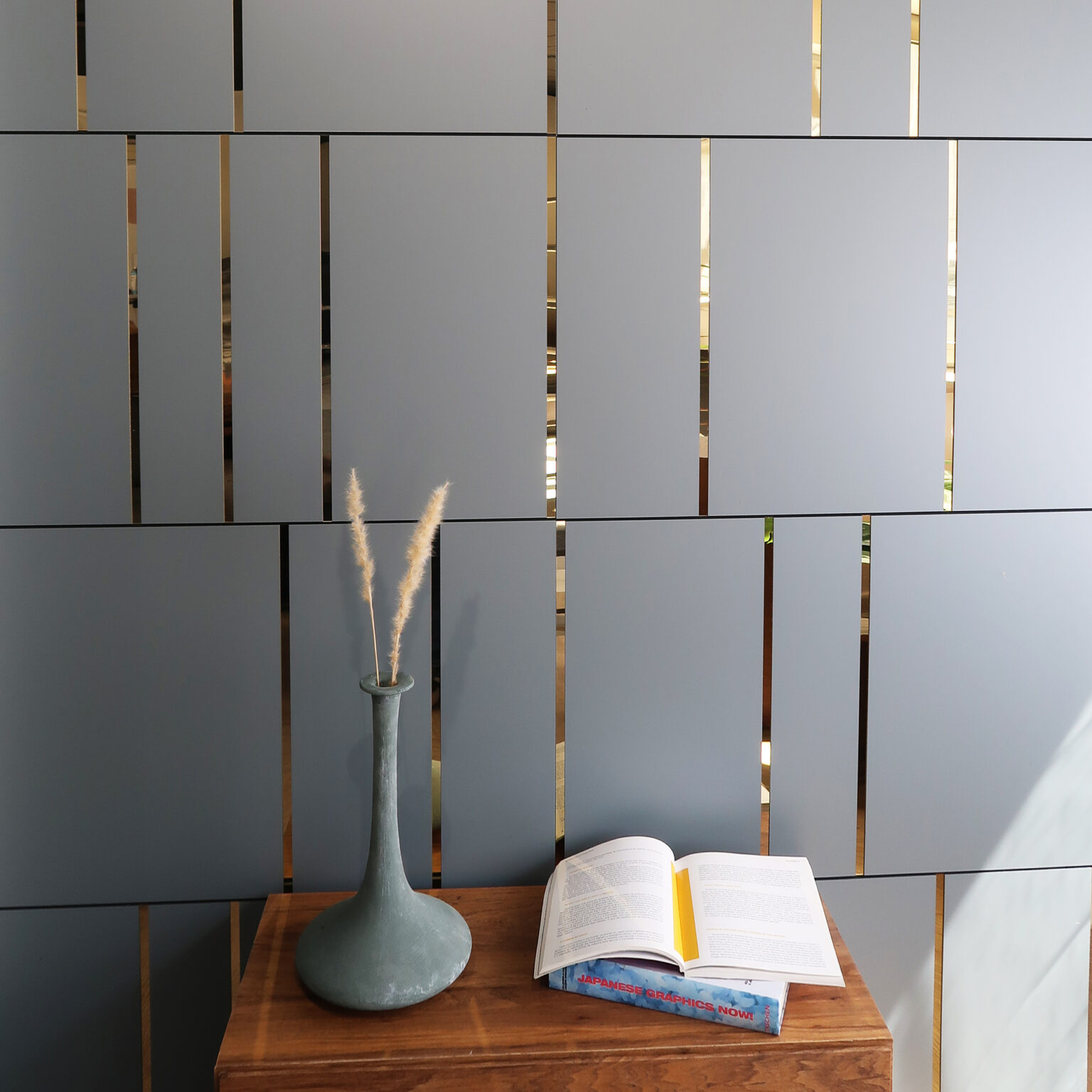 Wooden table with a tall gray vase holding dried grass and an open magazine, set against a gray slatted wall with gold gaps.