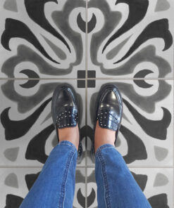 Top-down view of a person in blue jeans and black patent loafers standing on gray tiled floor with bold black abstract patterns.