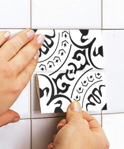 Hands press a black-and-white decorative patterned tile into place on a white tiled wall.