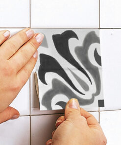 Person's hands apply a square decorative tile sticker with black and gray swirls to a white tiled wall.