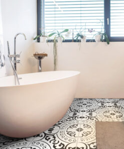 Modern bathroom with a white freestanding tub, chrome fixtures, and a row of potted plants on a windowsill above patterned black-and-white tiled flooring.