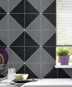 Black tiled kitchen wall with white geometric diamond patterns, a dish rack with plates and a glass on the counter, and a window with blinds and purple potted plants on the sill.