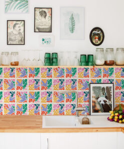 Bright kitchen display: a colorful tiled backsplash, glassware on a shelf, and jars along a white counter beneath framed art panels.