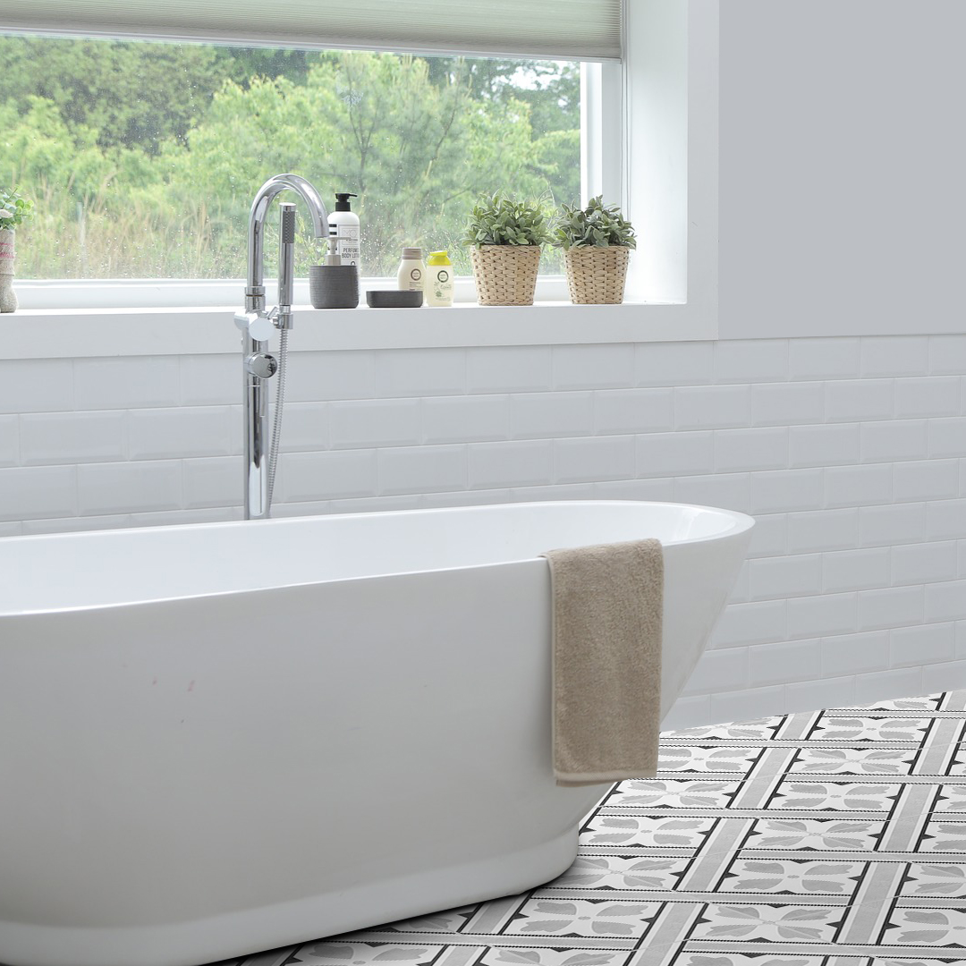 04 | Casadart | Vinis Autocolantes, Papel de Parede e Painéis Decorativos Bright bathroom featuring a white freestanding tub with a beige towel, chrome faucet, and a windowsill with small potted plants and toiletries; patterned floor tiles.