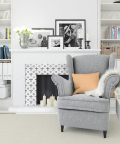 Gray armchair with an orange pillow sits in a white living room beside a decorated mantel and framed black‑and‑white photos.