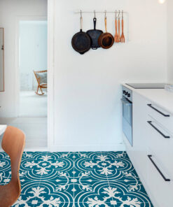 Bright white kitchen with bold teal patterned tile floors, white cabinets, and a wall rack holding cast-iron pans and wooden spoons. A doorway opens to a sunny room with a rocking chair in the background.