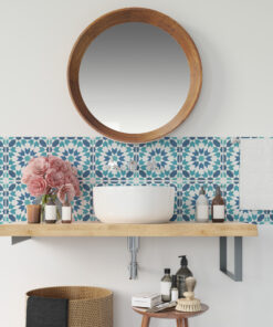Round wooden-framed mirror above a white sink with a blue geometric tile backsplash, pink flowers, and bathroom products on a wooden shelf and wicker basket nearby.