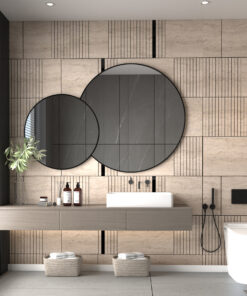 Bathroom with beige tiled wall, two overlapping black-framed round mirrors above a light wood vanity, plant and bottles on counter.