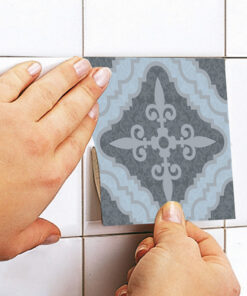 Hands press a decorative tile sticker onto a white tiled wall. The sticker has a gray and light blue damask pattern.