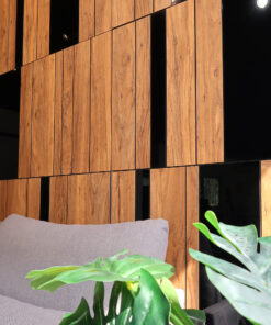 Wooden slat wall with narrow black gaps and a gray couch cushion in the foreground; green plant leaves in the foreground.