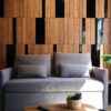 IMG_3677 copy | Casadart | Vinis Autocolantes, Papel de Parede e Painéis Decorativos Gray sofa with multiple cushions in front of a warm wooden panel wall; a wicker pot with a plant and a remote on a wooden table nearby.