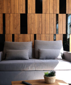 Gray sofa with multiple cushions in front of a warm wooden panel wall; a wicker pot with a plant and a remote on a wooden table nearby.