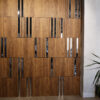 IMG_3969 copy | Casadart | Vinis Autocolantes, Papel de Parede e Painéis Decorativos Wood-paneled wall with alternating vertical black glass strips, creating a modern interior feature. Right side shows a light gray wall and plant leaves.
