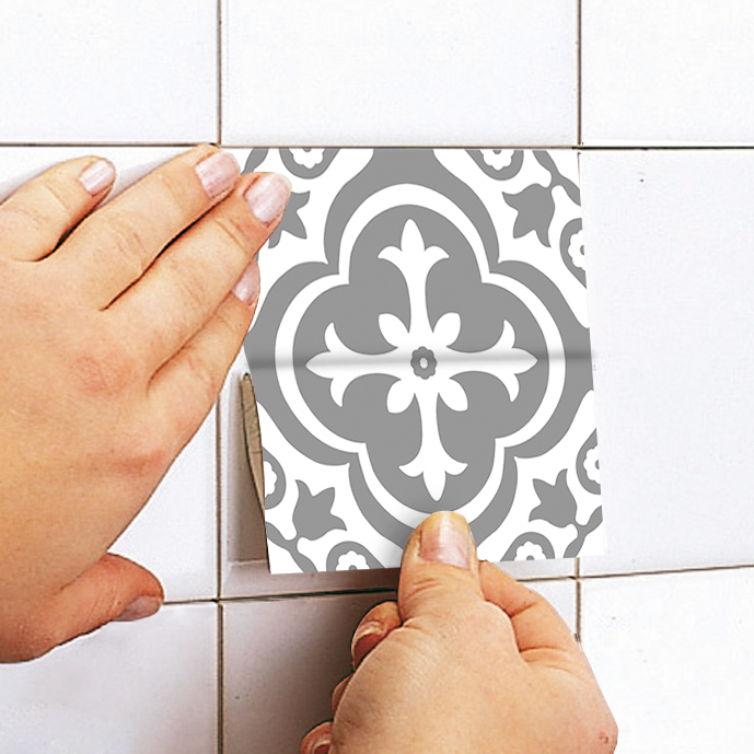 apply | Casadart | Vinis Autocolantes, Papel de Parede e Painéis Decorativos Close-up of hands applying a gray and white patterned tile sticker to a white tiled wall, aligning it with the grout lines.