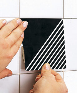 Hands press a decorative black tile with white diagonal stripes onto a white tiled wall.