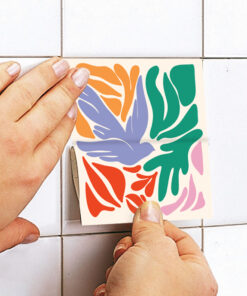 Two hands apply a vibrant multicolor abstract sticker to a white tiled wall, featuring orange, purple, green, red, and pink shapes.