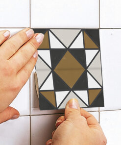 Close-up of hands installing a geometric mosaic tile on a white tiled wall; the tile has black, white, gray, and gold triangles forming a central diamond pattern.