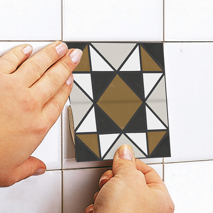 apply copy | Casadart | Vinis Autocolantes, Papel de Parede e Painéis Decorativos Close-up of hands installing a geometric mosaic tile on a white tiled wall; the tile has black, white, gray, and gold triangles forming a central diamond pattern.
