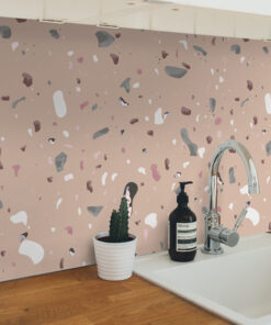 A peach terrazzo wall behind a white countertop with a plant in a white pot and a dark bottle beside a chrome faucet.