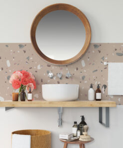 Round wooden-framed mirror above a white vessel sink in a terrazzo-tiled bathroom, with pink flowers and assorted grooming bottles nearby.